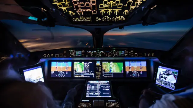 A imagem mostra o interior de uma cabine de comando moderna durante um voo ao amanhecer ou entardecer. Os painéis de instrumentos e telas digitais iluminam o cockpit com tons de azul, verde e amarelo, exibindo informações de navegação, altitude e sistemas da aeronave. Em primeiro plano, é possível ver parcialmente dois tripulantes sentados diante dos controles. Pela ampla janela frontal, observa-se o horizonte com o céu em tons azulados e alaranjados acima de uma camada de nuvens, criando uma atmosfera tecnológica e ao mesmo tempo contemplativa.