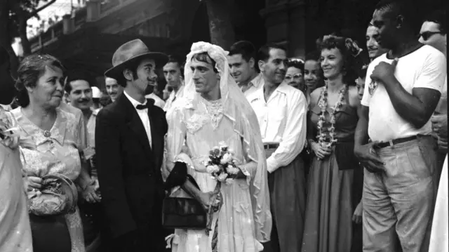Fotografia em preto e branco tirada no carnaval de rua do Rio de 1954 mostra ao centro um casal de noivos, com o homem fantansiado de noiva e a mulher, de noivo, e várias pessoas sorridentes em volta