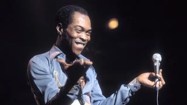 Nigerian singer Fela Kuti, wear pale blue long-sleeved shirt wit bits of yellow and pink embroidery, e dey smile and hold up im hands toward di audience at Vredenburg in Utrecht, Netherlands on 3 November 1988