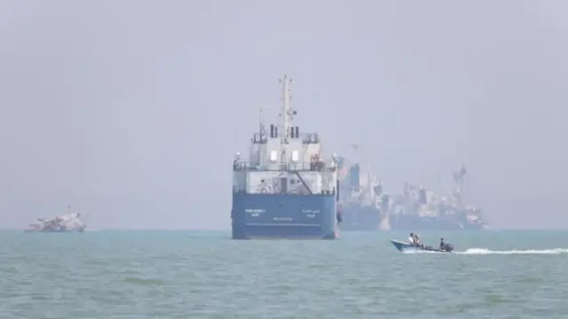 Denizden geçen önde bir tanker arkasında belirsiz iki gemi daha var. Öndekinin sağ açığında insanlı küçük bir tekne görünüyor. 