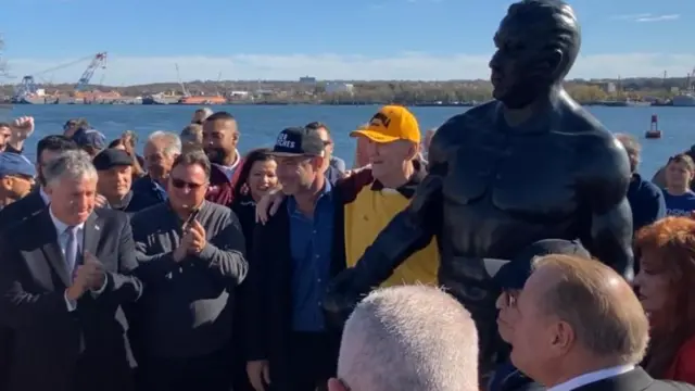 چاک وپنر (با کلاه زرد) در کنار مجسمه‌ای که به افتخارش در نیو جرسی نصب شده، ژست گرفته است