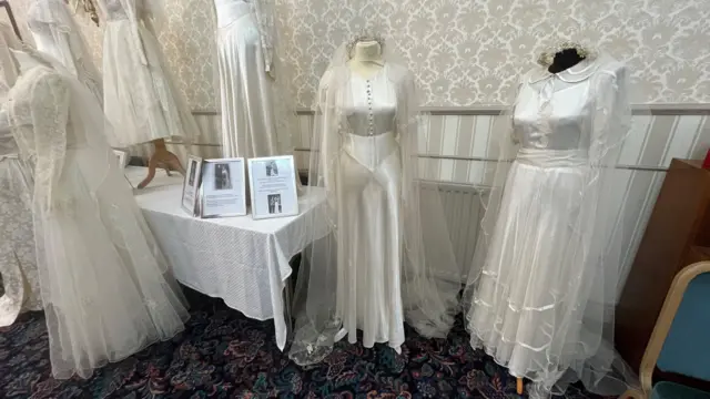 Біла шовкова весільна сукня з ґудзиками зверху та довгою фатою. Сукня з довгими рукавами та облягає талію.