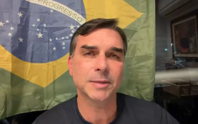 Flávio Bolsonaro falando para a câmera com uma bandeira do Brasil ao fundo