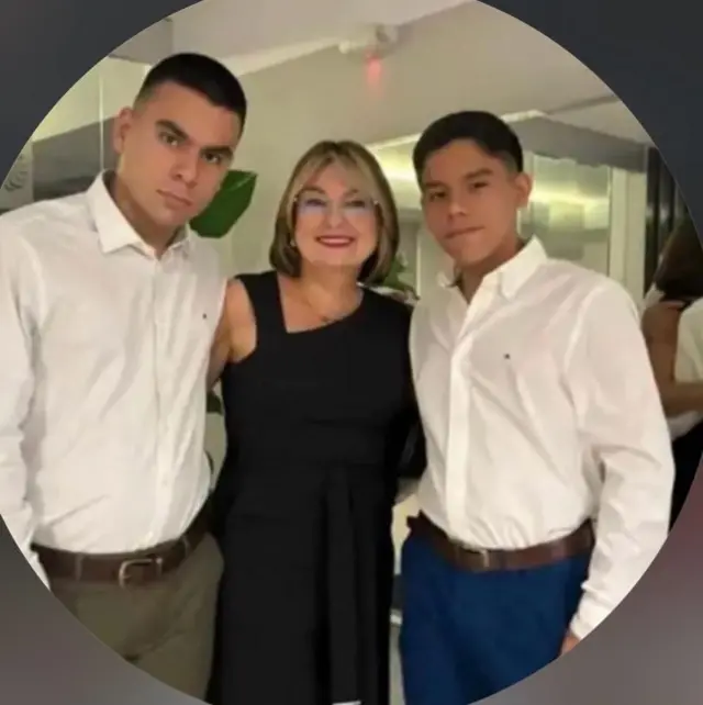 Al lado izquierdo, Santiago, con una camisa blanca y un pantalón verde, abraza a su madre, quien está en el centro. María Lourdes, sonríe, lleva lentes y un vestido negro. Al lado derecho, Gonzalo, vestido con una camisa blanca y un pantalón azul, también la abraza.