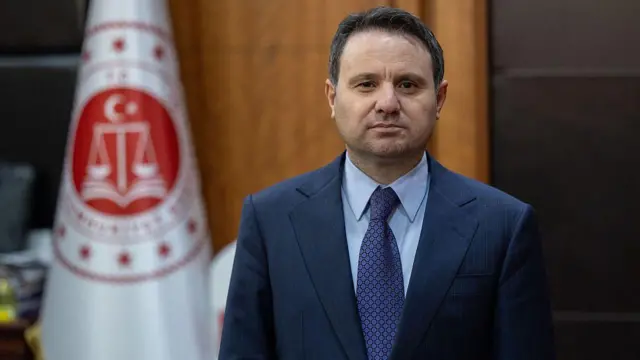 Akın Gürlek, Adalet Bakanlığı bayrağının önünde, ayakta poz veriyor. 