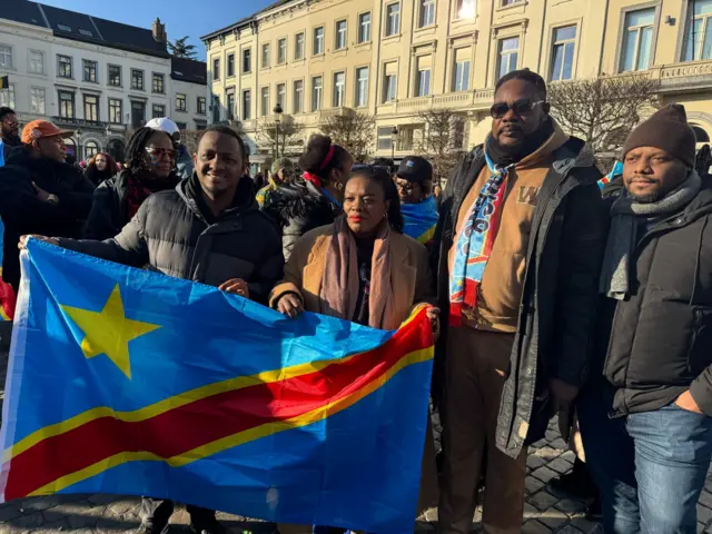 Lydia Mutyebele Ngoi hagati y’abanyekongo babiri imbere n'abandi inyuma afise ibendera rya Congo mu myiyerekano yo kwiyamiriza intambara mu buseruko bwa Congo imbere y’inama nshingamateka y’ibihugu vya Bulaya mu ntango z’ukwezi kwa gatatu mu 2025, i Bruxelles, Belgique.