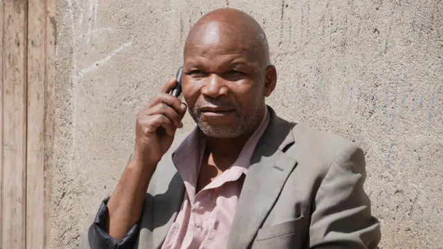 Umugabo wambaye ishati ijya kuba iroza ryerurutse n'ikositimu y'ibara ry'ikijuju, afite telefone ku gutwi. Inyuma ye hari urukuta rw'inzu n'umuryango w'inzu uboneka igice. 