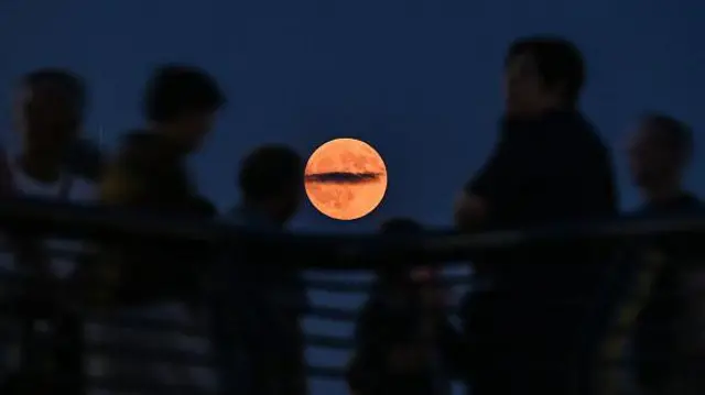 Várias pessoas olhando para a Lua de Sangue, que tem uma cor laranja claro, no centro da foto.