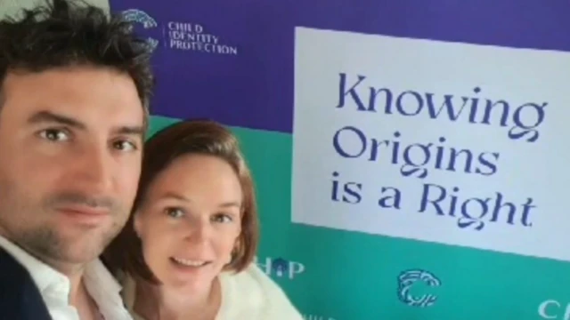 Audrey ve Arthur, üzerinde “Kökenleri Bilmek Bir Haktır” mesajı bulunan ve çocuk kimliği savunuculuğuyla ilişkili logolar içeren büyük bir kampanya pankartının önünde yan yana duruyor.