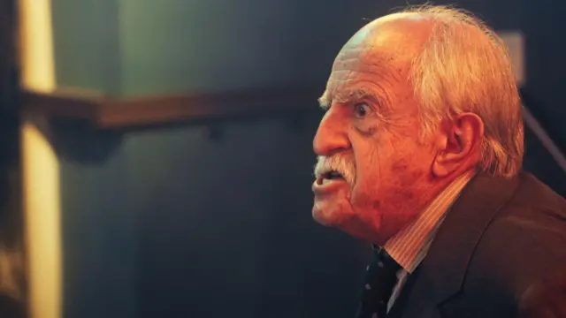 Ary Fontoura, de pele clara, com cabelos brancos ralos e bigode espesso, aparece de perfil com a boca aberta, como se estivesse discutindo. Ele veste um terno escuro com camisa listrada e gravata, em um ambiente de iluminação quente que destaca as rugas e a tensão em seu rosto.