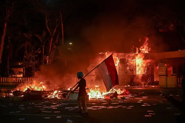 Seorang pengunjuk rasa berjalan dengan bendera Indonesia di depan markas polisi yang dibakar dan dijarah selama demonstrasi di Surabaya pada 31 Agustus 2025.