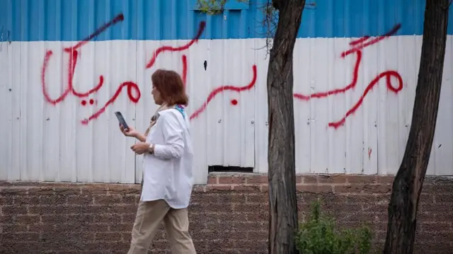 زنی بدون حجاب اجباری از جلوی دیواری که روی آن نوشته «مرگ بر آمریکا» عبور می‌کند