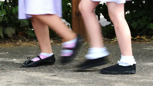 Se ven dos pares de piernas de niños caminando por la acera. Llevan calcetines cortos y zapatos escolares negros. Se ve la parte inferior de las faldas de cuadros vichy del uniforme escolar justo por encima de las rodillas.