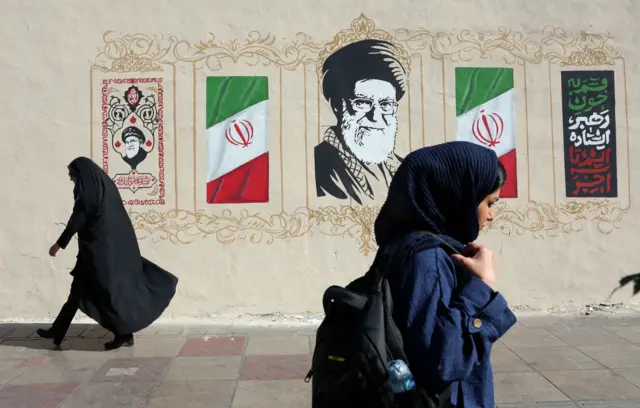 Dos mujeres con velo caminan por la acera frente a un mural que tiene banderas iraníes, caligrafía decorativa y un gran retrato de Alí Jamenei. Una mujer lleva una túnica negra, mientras que la otra viste un abrigo azul oscuro y lleva una mochila.