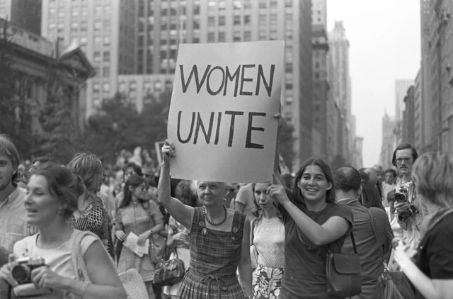 Una mujer mayor y una joven sostienen un cartel que dice "Mujeres Unidas" mientras marchan, con más mujeres, por la Quinta Avenida en la ciudad de Nueva York