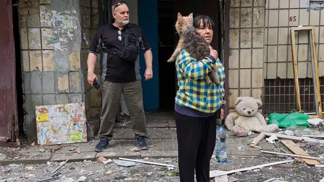 Zniszczone podwórko, na którym stoi para ludzi, kobieta trzyma kota, dookoła leży gruz.
