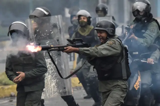 Un miembro de la Guardia Nacional dispara su escopeta contra manifestantes durante enfrentamientos. Está rodeado de otros miembros de la Guardia Nacional. Todos visten de color verde militar, con chaleco antibalas, casco. En muchos casos llevan la cara tapada. otros llevan un escudo transparente de protección. El militar que está en el centro sostiene un arma larga en sus manos. de la boca del arma se ve fuego.