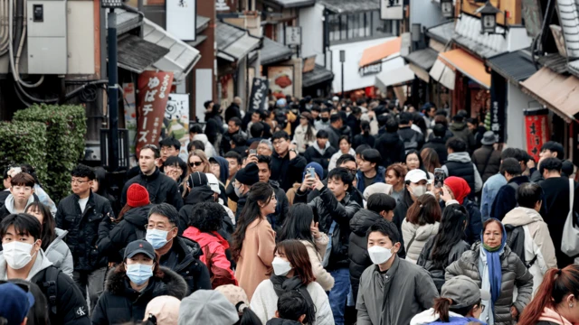 拥挤的街道上，游客人潮熙攘，手持手机、并佩戴口罩。