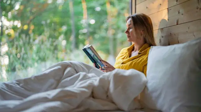 Jovem sentada, lendo em uma cama junto à natureza