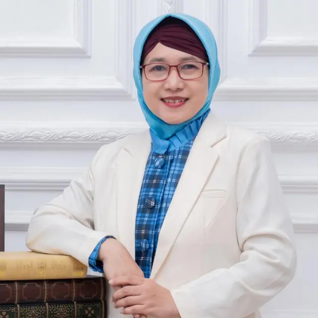 Profesor Zullies Ikawati dengan blus putih dan dalaman kemeja kota biru yang seragam dengan hijabnya sedang menatap ke arah kamera dengan gaya santai.