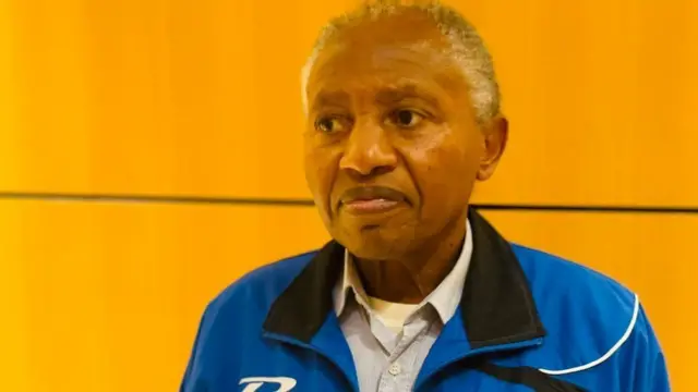 Umugabo w'umusatsi w'imvi, yambaye ishati y'ubururu bwerurutse, arengejeko ijaketi y'ibara ry'ubururu