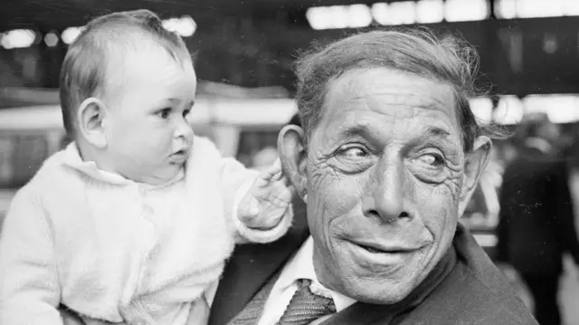 Un bébé vêtu d'un cardigan blanc effleure l'oreille de l'homme qui le porte. L'homme, vêtu d'une veste et d'une cravate, détourne le regard de l'enfant. Tous deux font partie des habitants de l'île de Tristan da Cunha, récemment arrivés au Royaume-Uni après l'évacuation volcanique de 1961.