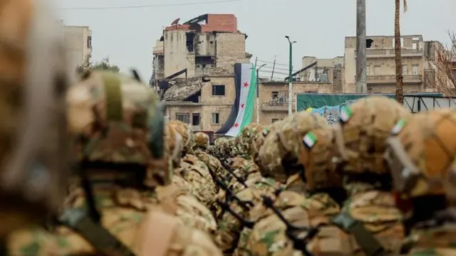 Kamuflajlı askerler Suriye bayrağı asılı yarısı yıkılmış bir binaya doğru ilerliyor