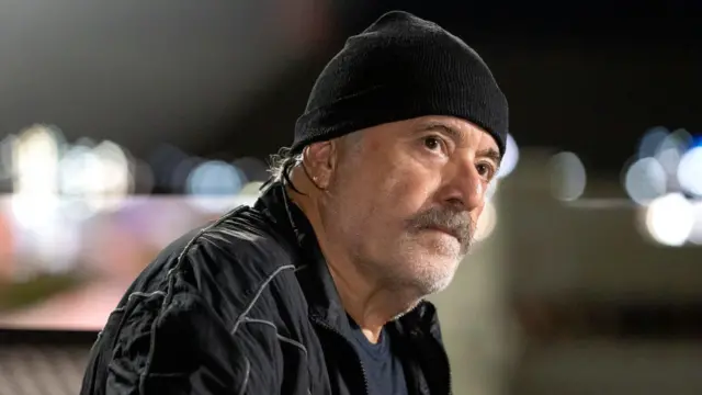 Tony Ramos usa uma jaqueta e um gorro pretos. Ele tem barba grisalha curta e expressão séria, com o olhar direcionado para algo fora do enquadramento, sugerindo concentração ou preocupação. O fundo está desfocado, com luzes que criam um efeito de bokeh, indicando um ambiente urbano à noite.