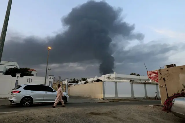 رجل يسير بجوار مجمع سكني، بينما يتصاعد عمود كثيف من الدخان الأسود عقب انفجار في المنطقة الصناعية بالفجيرة في 3 مارس/آذار 2026، ويرتدي الرجل سرولاً وقميصاً، وبيده حقيبة بلاستيكية 