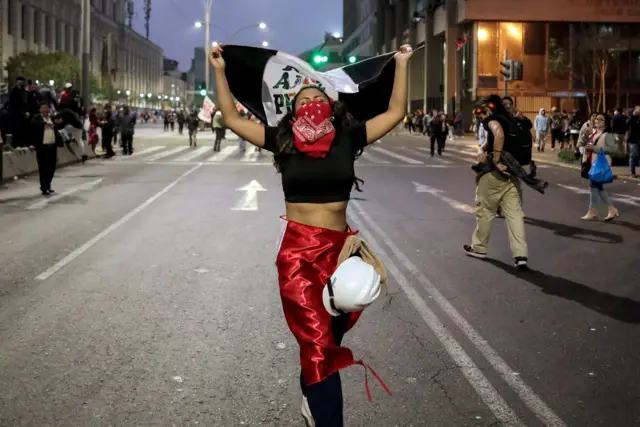 Una manifestante caminando por la calle con un pañuelo rojo en la cara, carga una bandera blanca y negra y un casco blanco le cuelga en la cintura