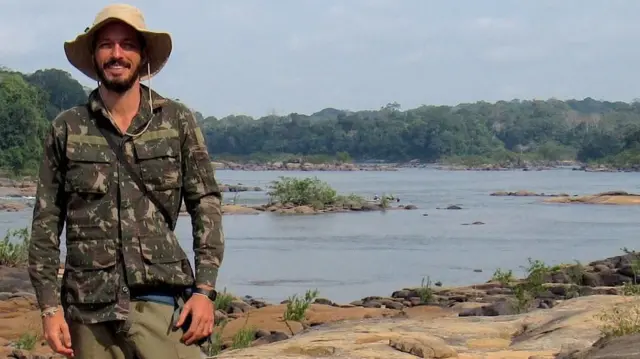 Rodrigo em pé, sorrindo, em frente a aparentemente uma lagoa ou rio