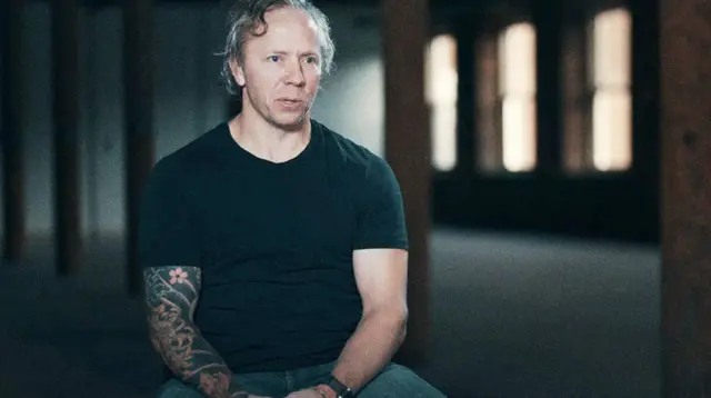 Портрет Грега Сквайра - седого мужчины, он сидит в полутемном помещении с деревянными колоннами и окнами, освещенными мягким светом. На нем чёрная футболка, видна татуировка на правой руке.