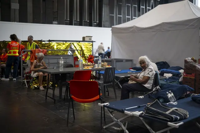 En segundo plano, a la derecha, una mujer mayor, con pelo blanco, mira a la cámara. Está sentada en una silla, rodeada de otras sillas de distintos tipis en mitad de un centro de refugio improvisado. Al fondo hay una carpa blanca. A la izquierda, al fondo, otra señora mayor con pelo blanco está sentada frente a una mesa. Más al fondo, dos voluntarios del refugio visten chalecos reflectantes.