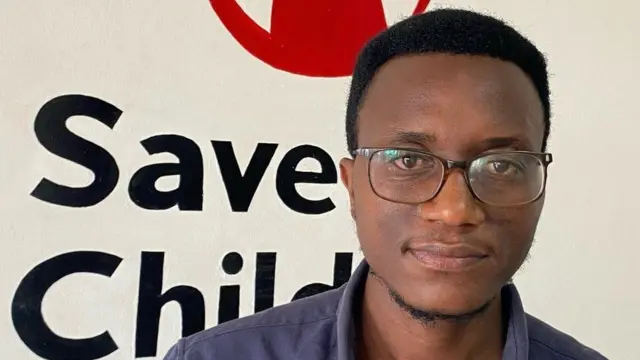 Umugabo wambaye indorerwamo arimo kureba muri 'camera', inyuma ye hari ikirango cya 'Save the Children'