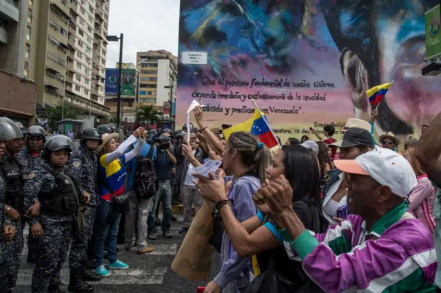 Акція протесту у Венесуелі. Зліва в ланцюзі стоять поліцейські в обмундируванні, поряд з ними дівчина, яка знімає право протестуючих. Вони щось скандують перед поліцією. На задньому плані мурував і житлові будинки