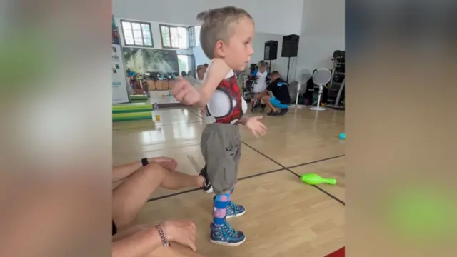 Edward en una sala de fisioterapia parado por sí solo. Tiene un soporte corporal y una mujer adulta está sentada atrás de él