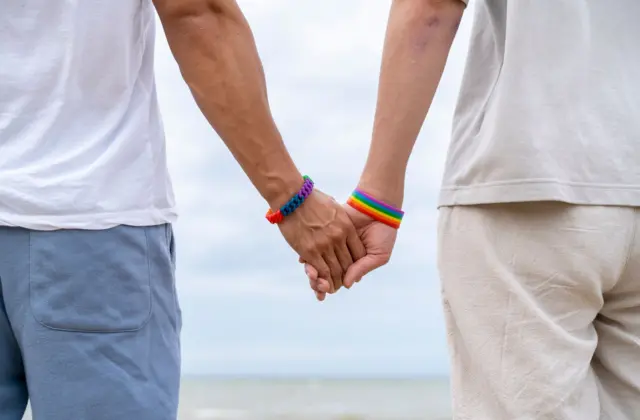 
Um casal homossexual de mãos dadas em frente à praia.
