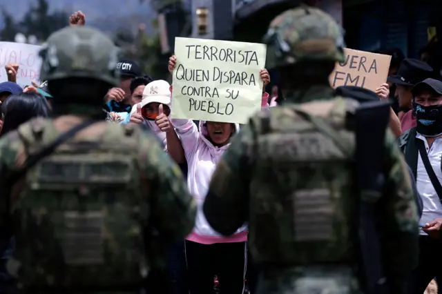 Mulher levanta cartaz que diz 'terrorista é quem dispara contra seu povo'; na frente dela, militares fortemente armados