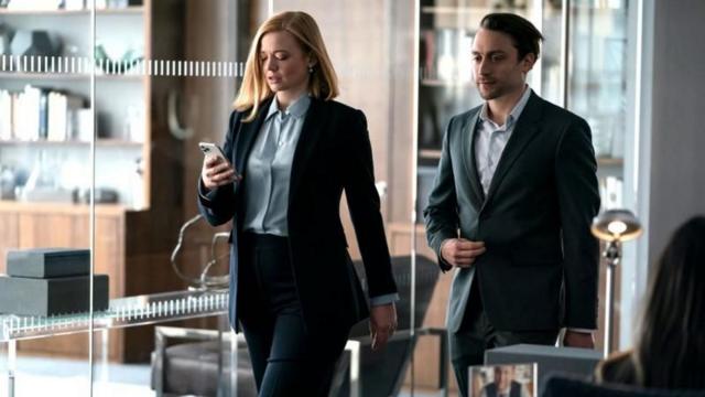 Atriz e ator caminhando em escritório em cena da série