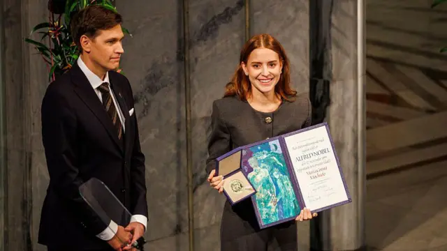 Ana Corina Sosa Machado, hija de María Corina, sostiene en sus manos la medalla del nobel de la paz y un certificado. sonríe. viste ropa gris, tiene cabello hasta los hombros. A su lado está Jorgen Watne Frydnes, quien sonríe. Viste un traje y corbata. 