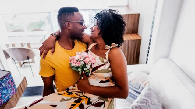Casal sorridente abraçado ao lado de um sofá, olhando um para o outro; ela segura um pequeno buquê de flores em uma sala iluminada.