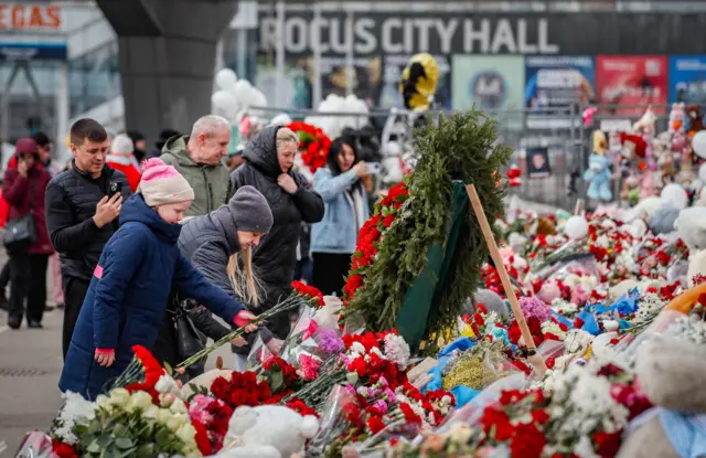 People add flowers to large display of tributes, on front of a building labelled Crocus City Hall