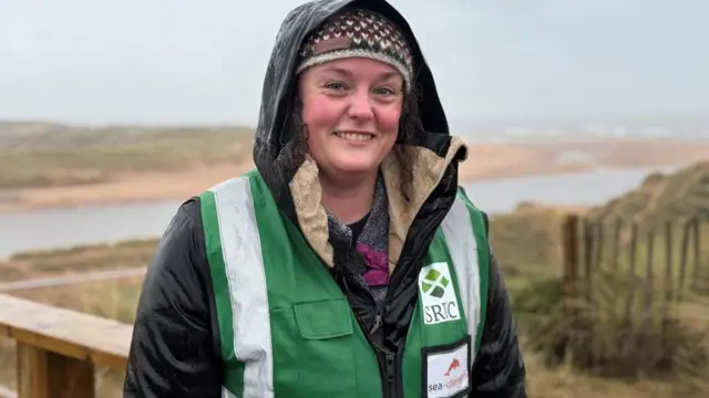 Claire Stainfield sonríe a la cámara con la playa de Newburgh en Aberdeenshire detrás de él, lleva una chaqueta y un gorro de punto de lana.