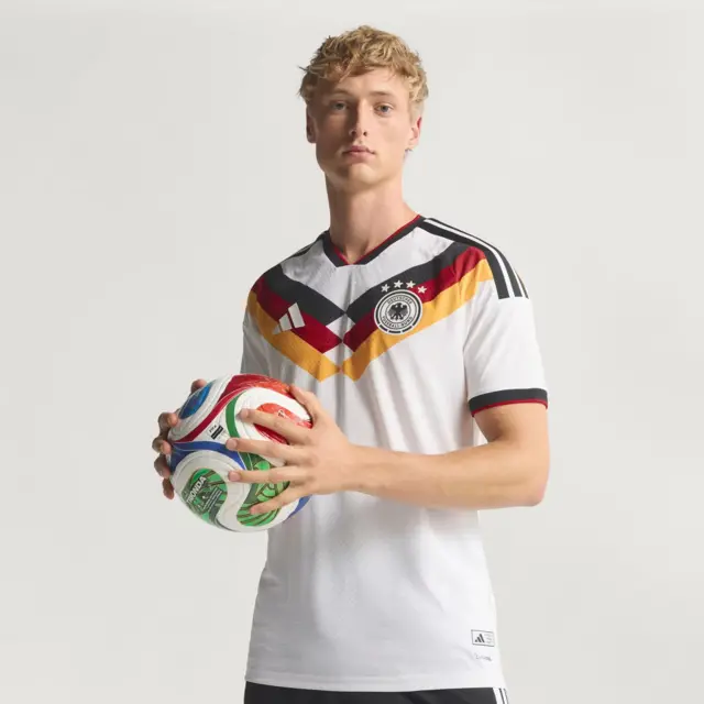 A imagem mostra um jovem segurando uma bola de futebol colorida com as duas mãos enquanto veste a camisa branca da seleção da Alemanha. O uniforme tem detalhes geométricos em preto, vermelho e amarelo no peito, remetendo às cores da bandeira alemã, além do escudo da federação do país com quatro estrelas e o logotipo da Adidas.