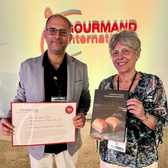 Gözlüklü bir erkek ile beyaz saçlı bir kadın kazandıkları ödülü ve yazdıkları kitabı ellerinde tutuyorlar. Keyifleri yerinde gözüküyor.