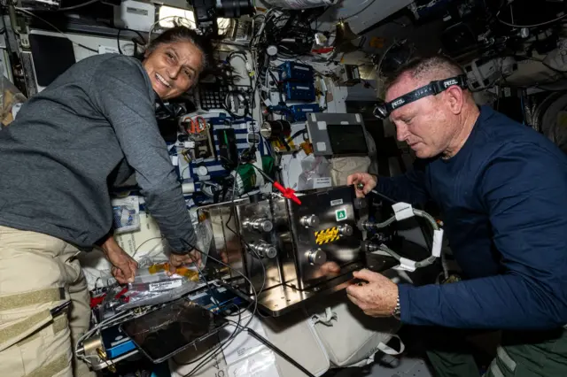 Butch e Suni na ISS - ambos estão usando roupas casuais e trabalhando em um equipamento que parece ser complicado, com muitos fios e consoles visíveis. Suni está inclinada sobre um equipamento e sorrindo, olhando por cima do ombro para algo. Butch está com uma lanterna na cabeça e concentrado em uma tarefa.