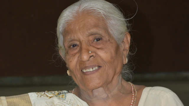A close-up of Meenakshi. She is looking at the camera and wearing white clothes. She is smiling, has grey hair and is wearing golden earrings. She also has a nose piercing. 