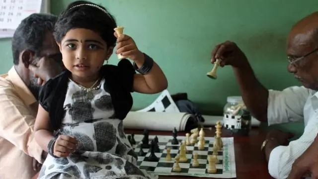 Una niña sosteniendo una pieza blanca. Al fondo, dos hombres juegan una partida de ajedrez