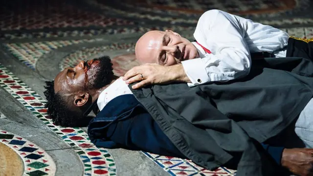  um homem negro de barba e terno está deitado no chão, com sangue no rosto, enquanto um homem branco e calvo de camisa se deita sobre seu peito