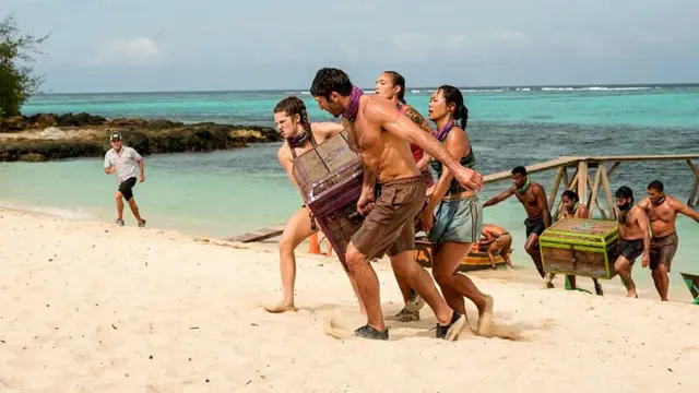 Escena de Survivor con participantes cargando cofres en la playa